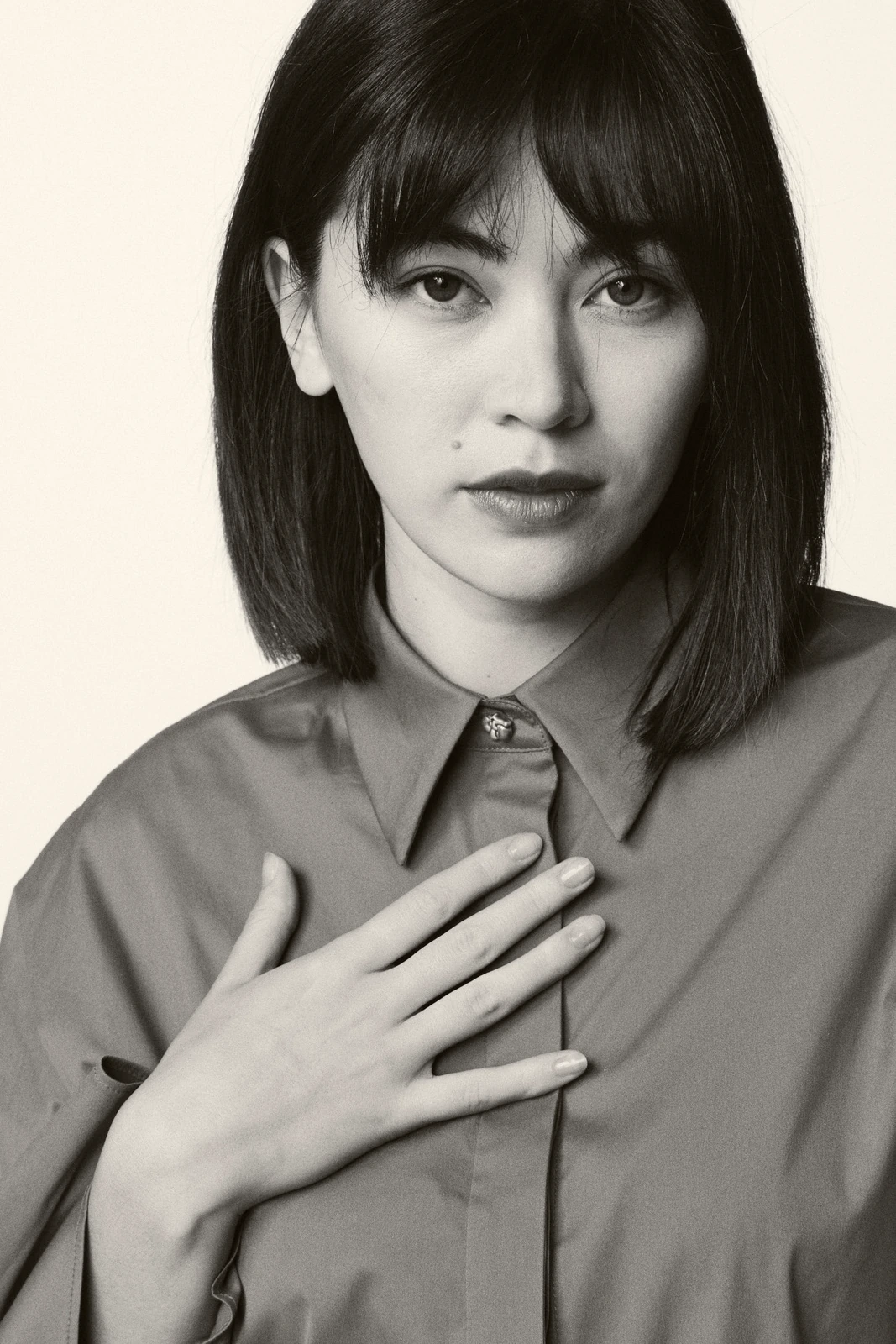 Actor Jessica Henwick, hand pressed to chest in an oversized shirt, direct gaze and bangs framing a cool, still.