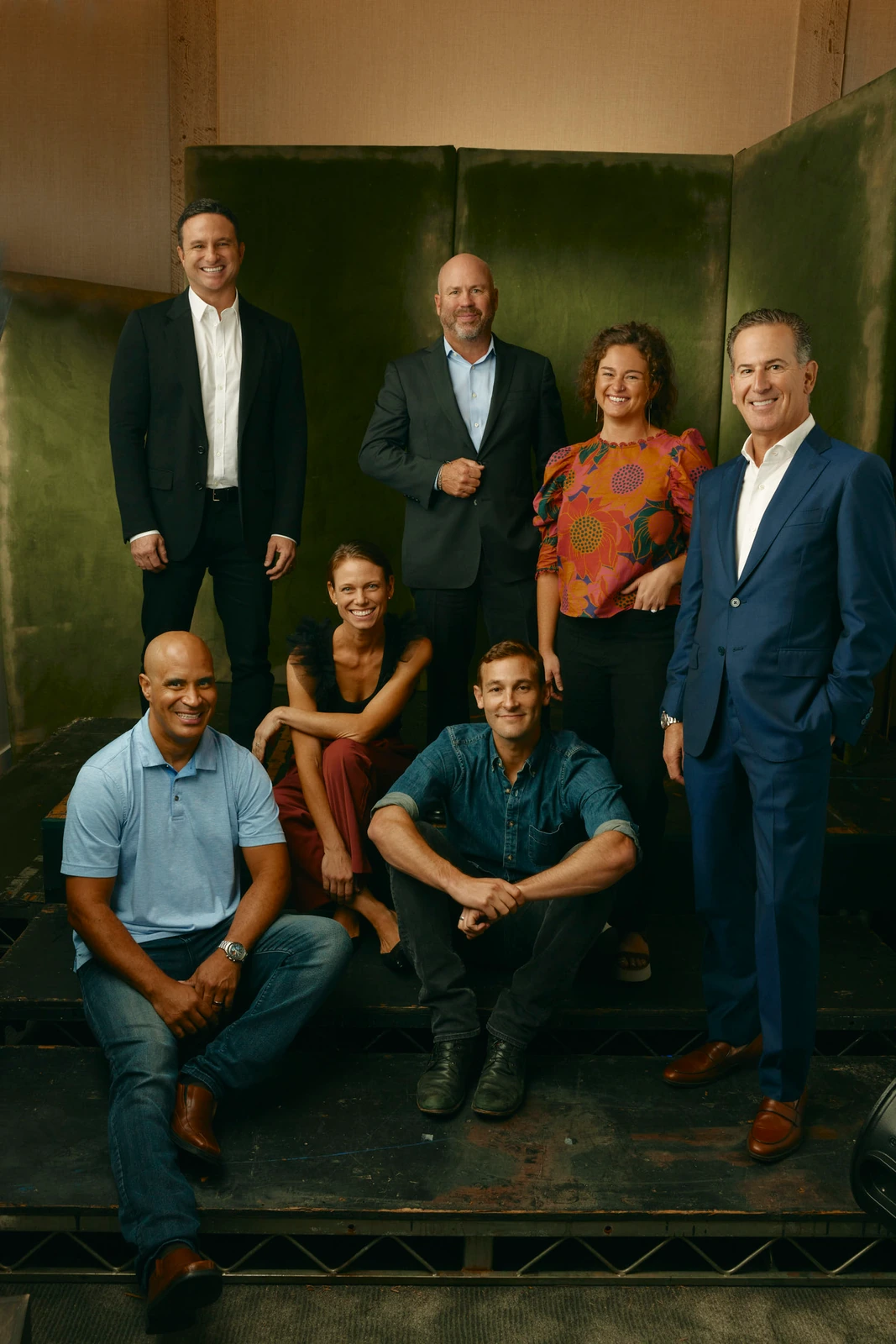 Group portrait of seven at Toronto Film Festival 2022, layered on worn green panels, warm light, relaxed mix of suits.