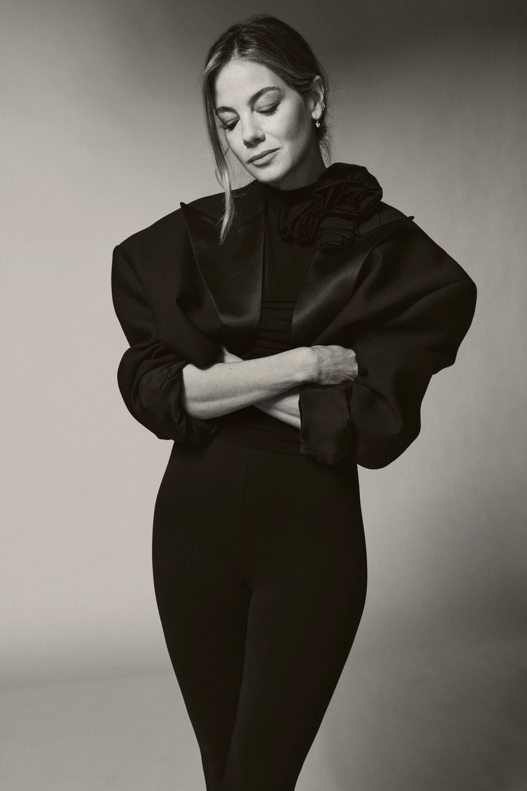Actor Michelle Monaghan, arms crossed in a sculptural black jacket with floral detail, gaze downward, soft light at TIFF.