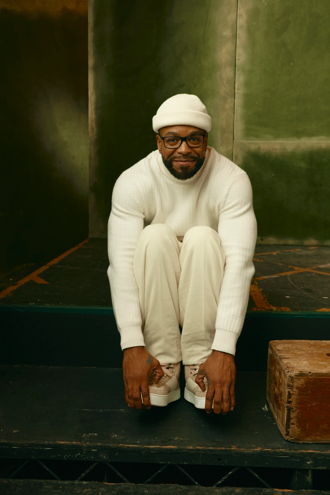 Actor Method Man seated on dark steps in head-to-toe cream — chunky knit, beanie, sneakers — against patinated green at TIFF.
