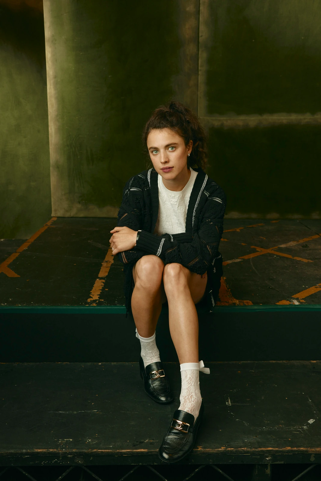 Actor Margaret Qualley seated on dark steps at TIFF 2022, black cardigan over white knit, lace socks and loafers, cool green.
