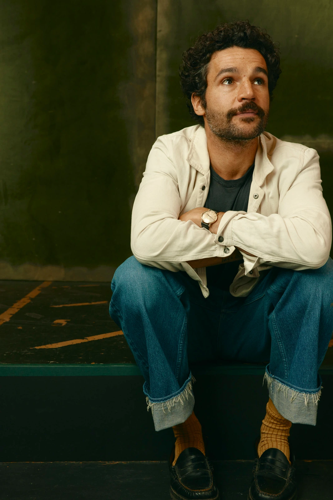 Actor Christopher Abbott seated arms crossed on dark steps, gazing upward in a white chore coat and cuffed jeans, warm olive.