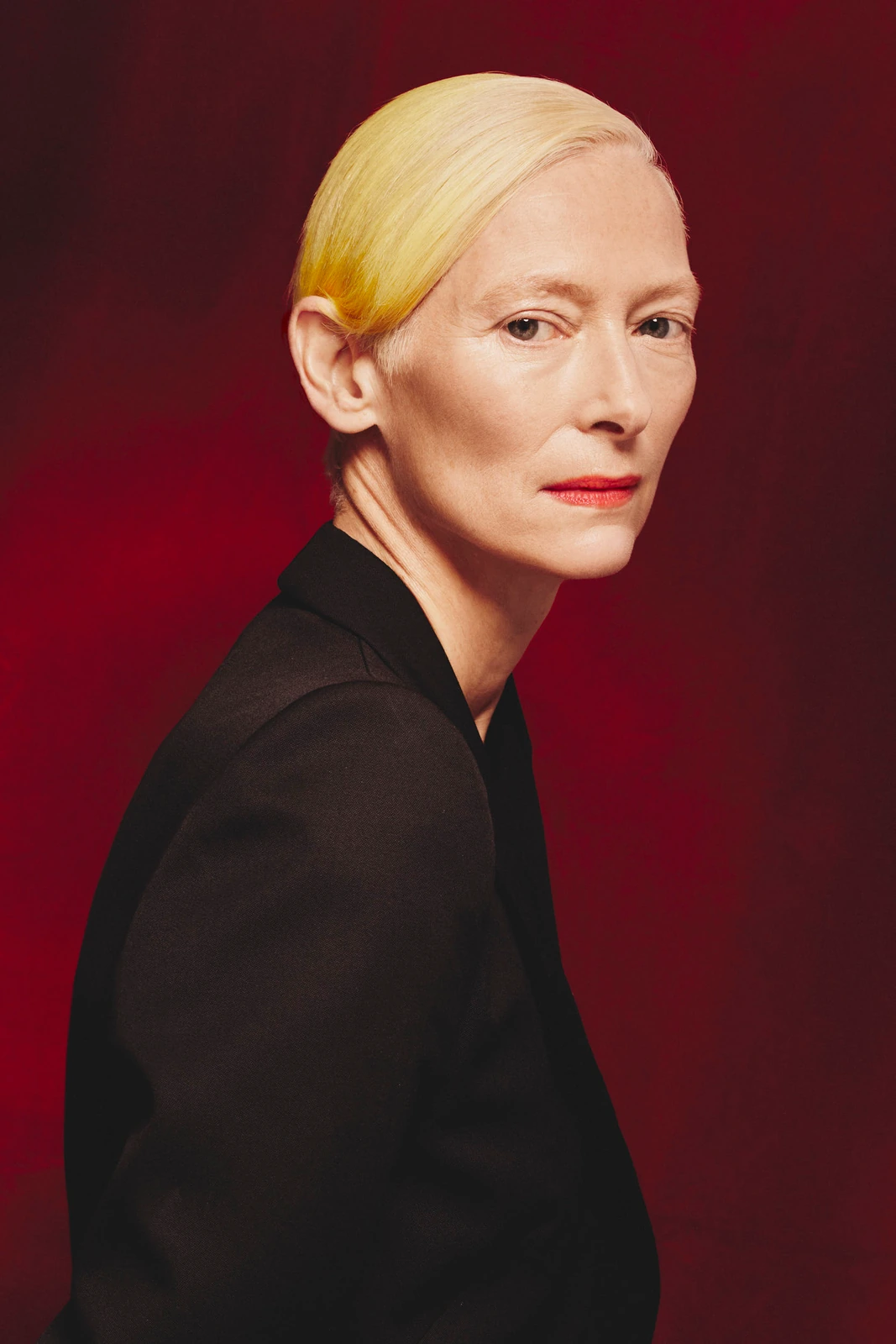 Actor Tilda Swinton, platinum hair slicked back, red lips, black blazer, gaze over shoulder against deep crimson — TIFF 2022.
