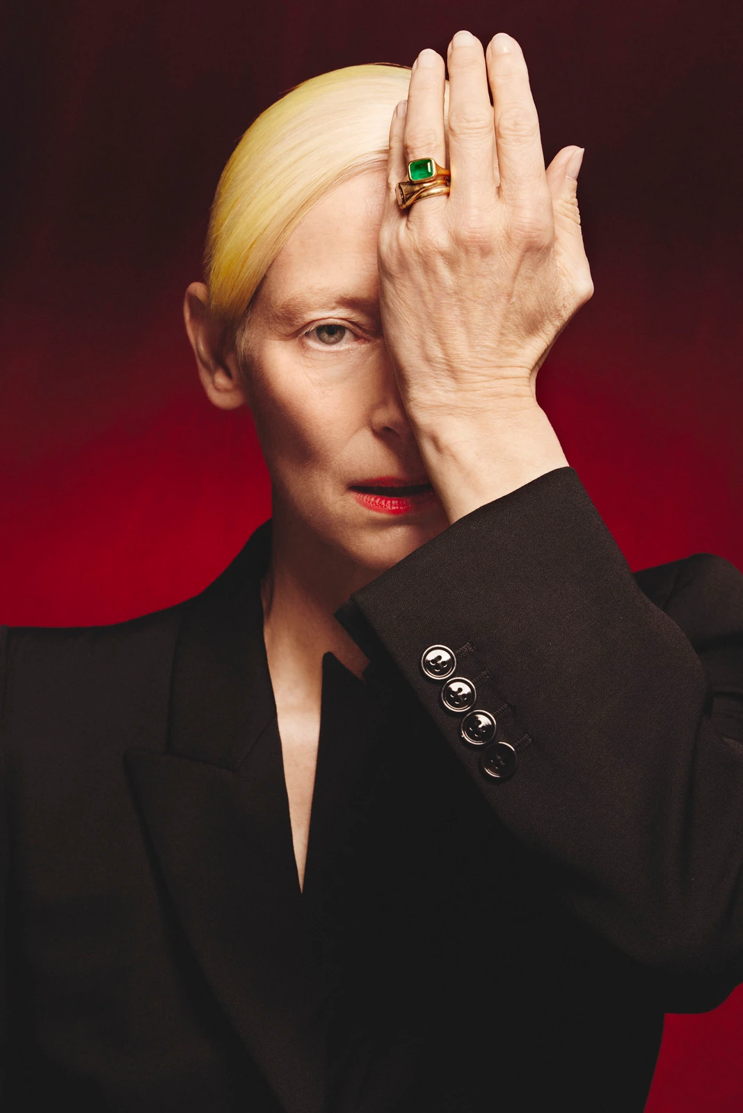 Actor Tilda Swinton, platinum hair slicked back, hand raised over one eye, emerald ring catching light against a deep.