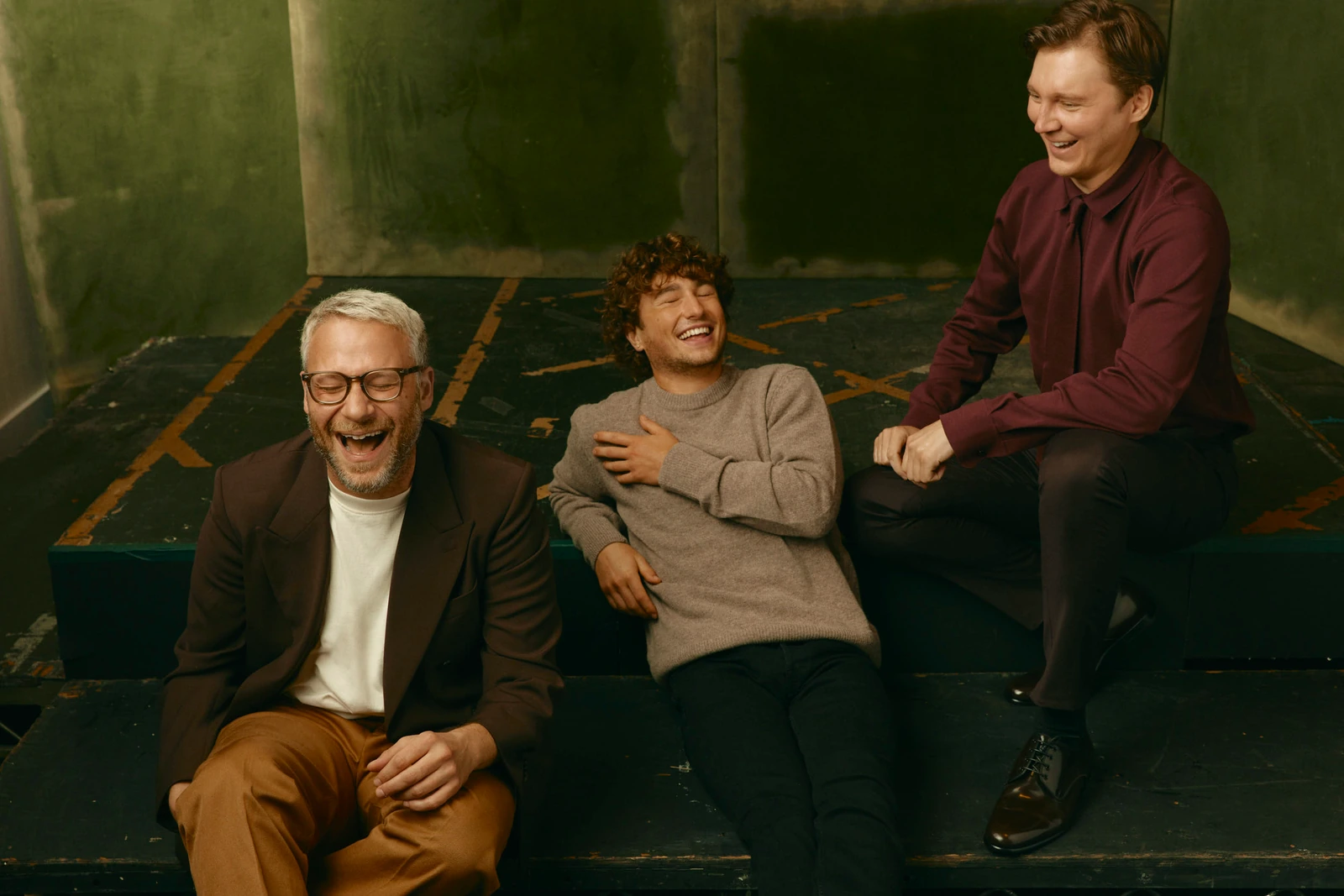 Actors Seth Rogen, Gabriel LaBelle, and Paul Dano laughing on a worn stage riser against a moody green backdrop, TIFF 2022.