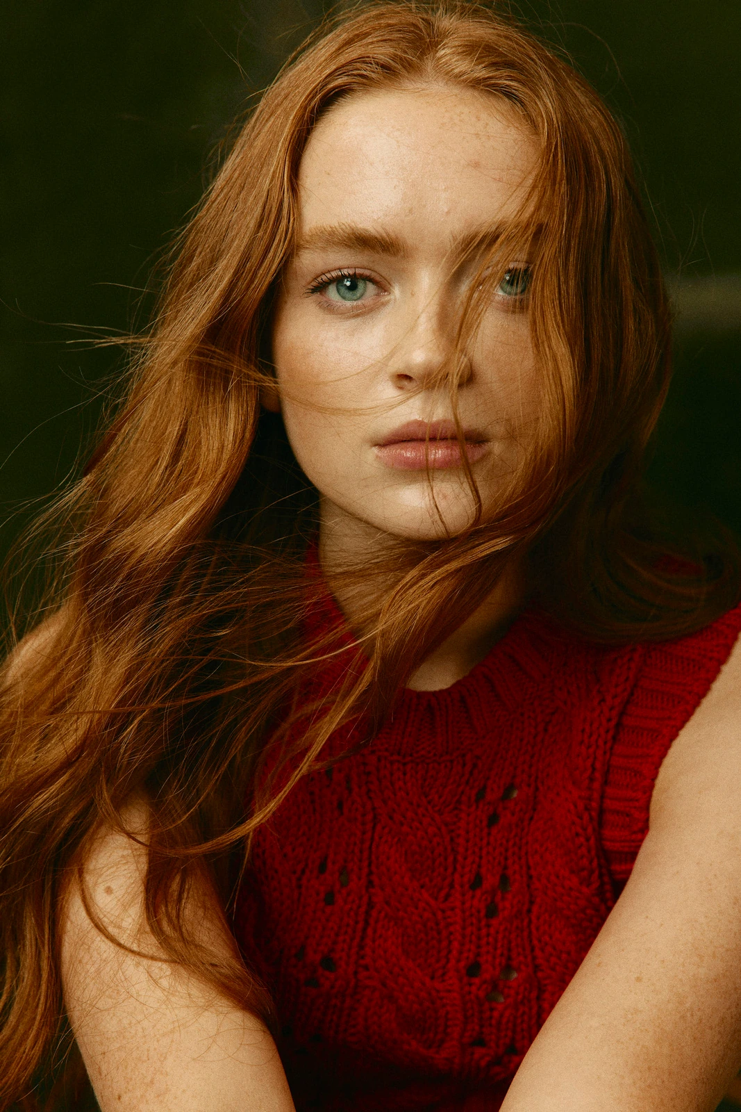 Actor Sadie Sink, copper hair windswept across her face, blue eyes steady, in a red cable-knit vest against deep green.