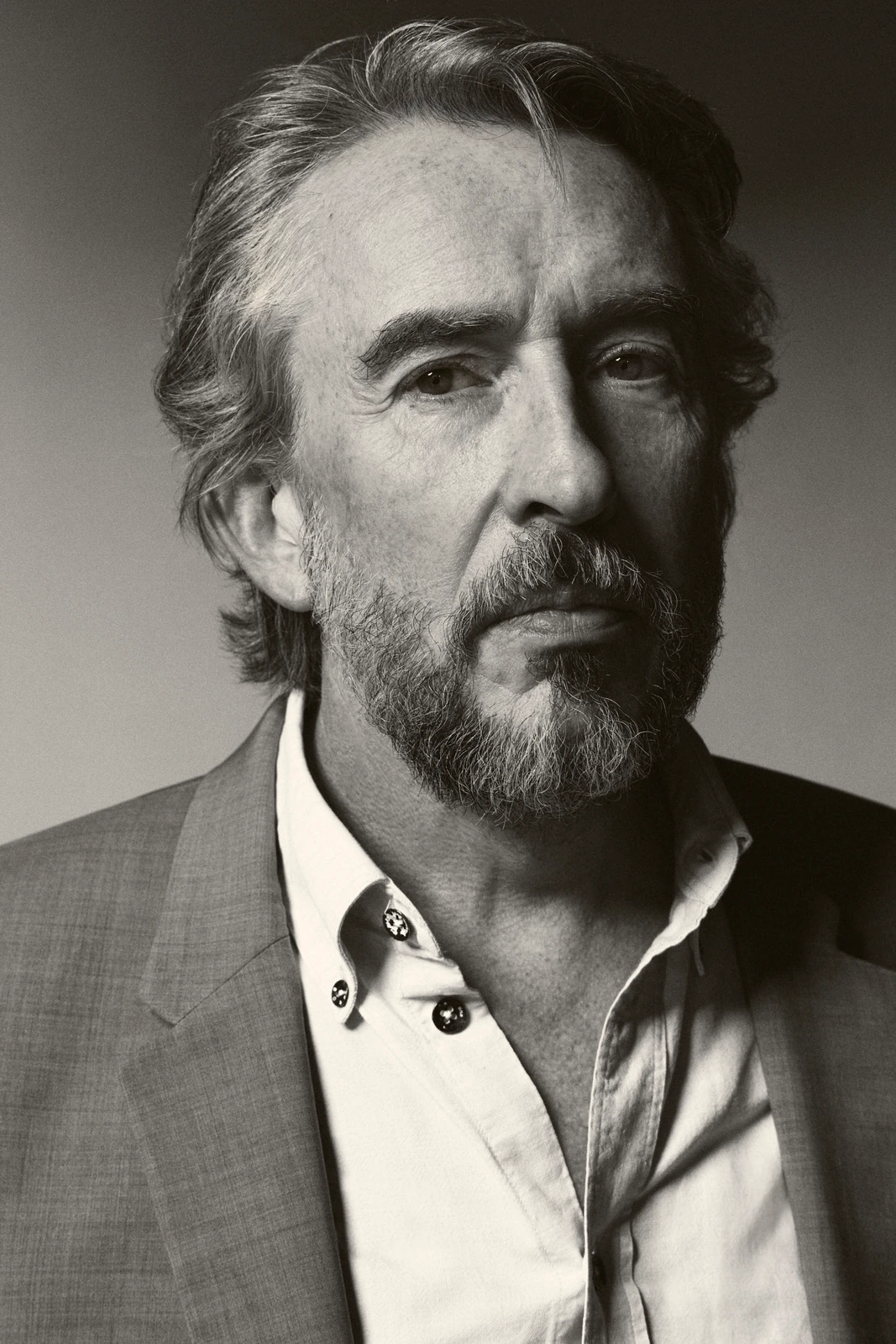 Actor Steve Coogan in a black-and-white studio portrait, grey blazer open at the collar, gaze distant and weathered.