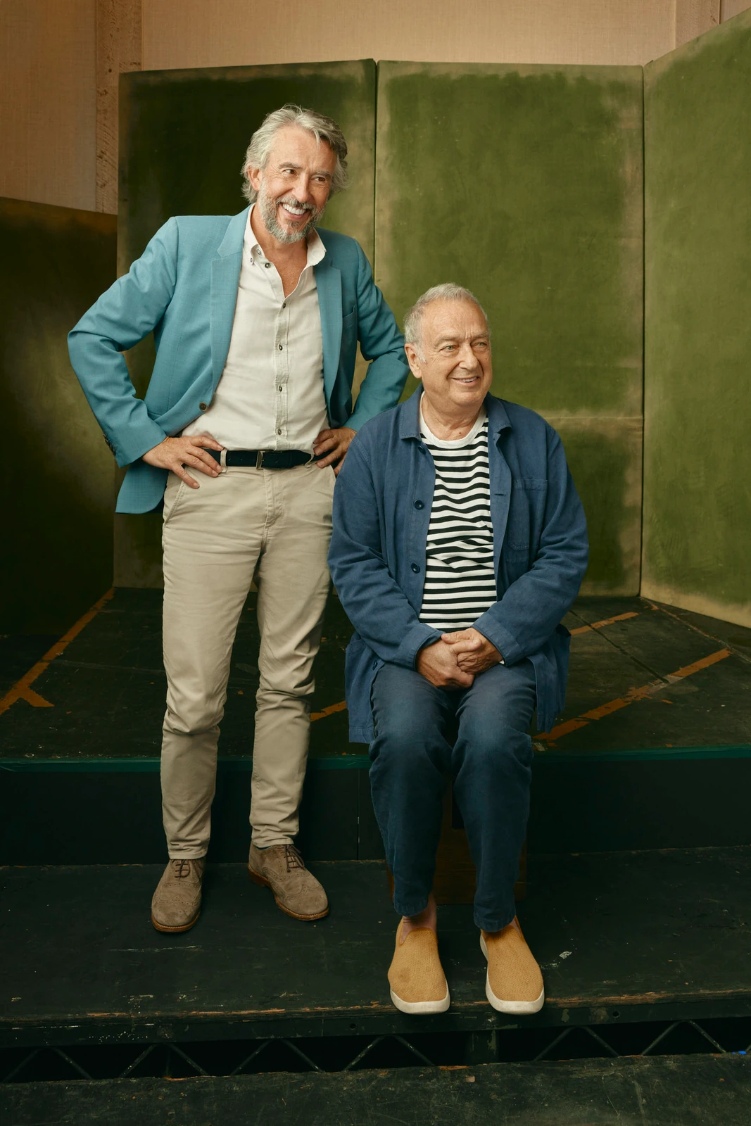 Actor Steve Coogan standing in a teal blazer beside director Stephen Frears seated in a navy jacket, warm green velvet.