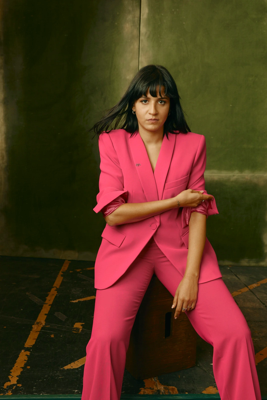 Actor Sara Mardini in a bold pink power suit, seated and direct-eyed against a moody olive-green wall at TIFF 2022.