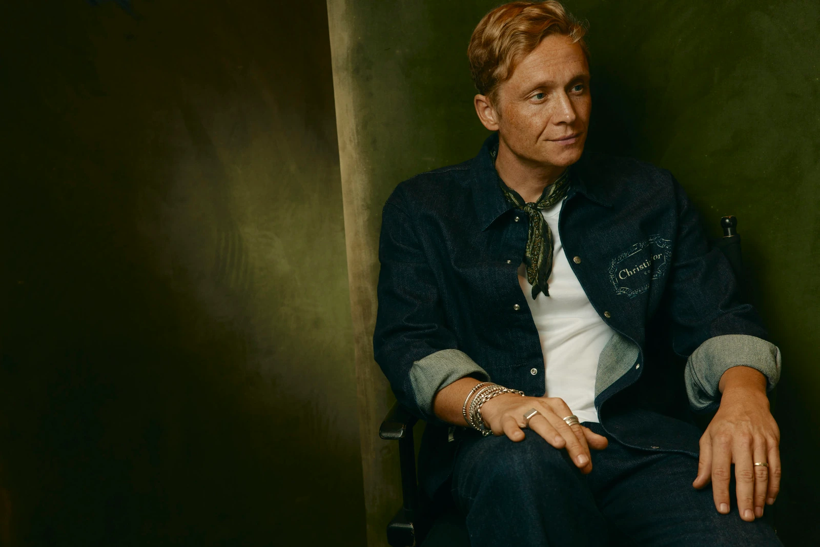 Actor Matthias Schweighofer seated in a chair, Dior denim jacket and olive neck scarf, stacked bracelets, warm green.
