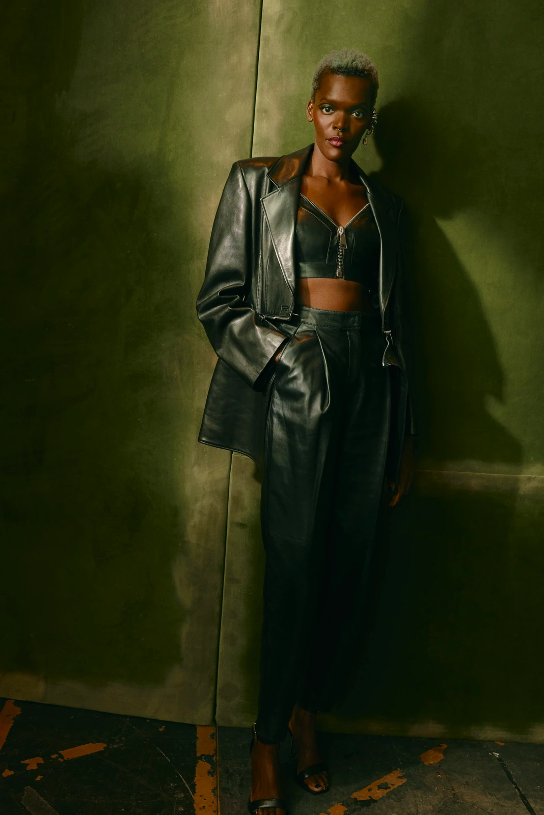 Actor Sheila Atim in head-to-toe black leather — oversized blazer, zip bralette, wide trousers — leaning against an olive.