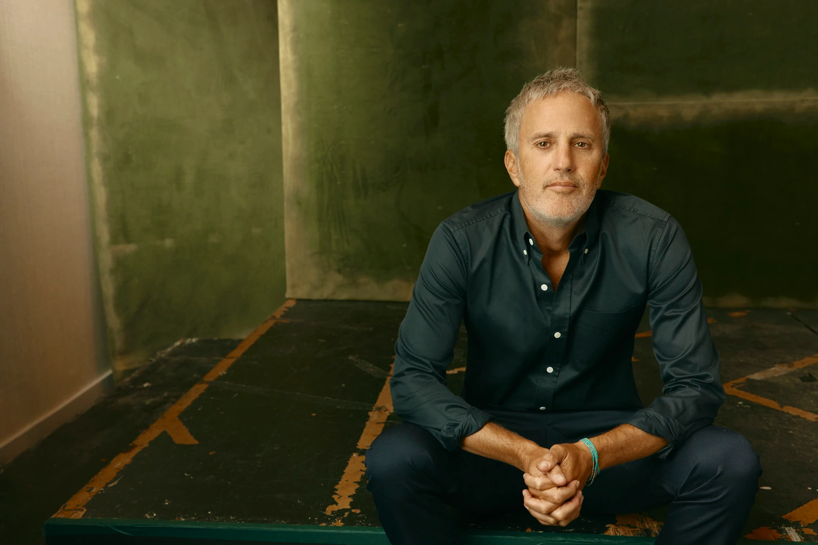 Actor Matt Smukler seated cross-legged on a worn stage floor, hands clasped, teal shirt, against a deep olive wall at TIFF.