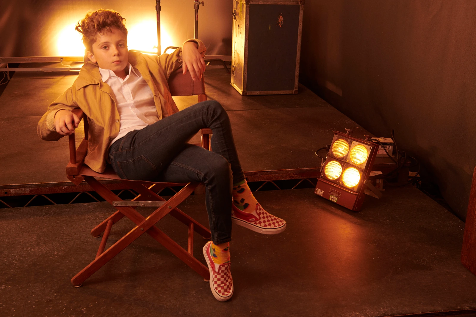 Actor Roman Griffin Davis slouched in a director's chair on a warmly lit stage, mustard jacket, checked Vans, quiet cool.