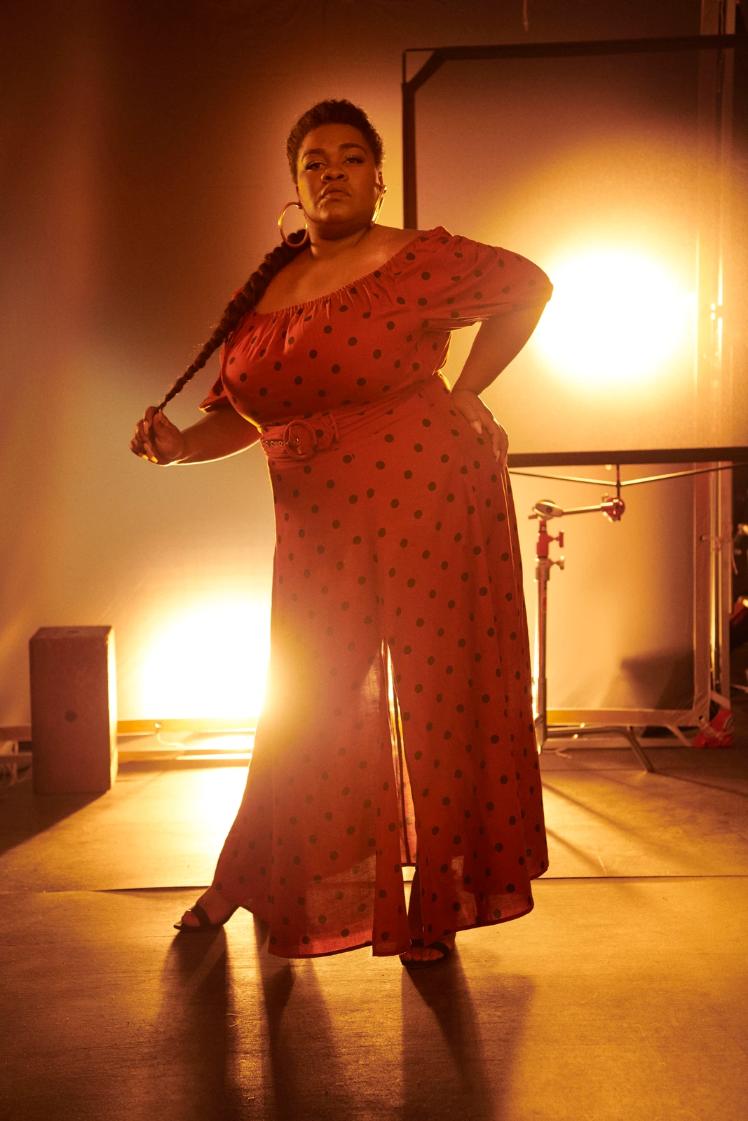 Actor Da'Vine Joy Randolph in a red polka-dot off-shoulder dress, hand on hip, braided hair in hand, backlit by warm amber.