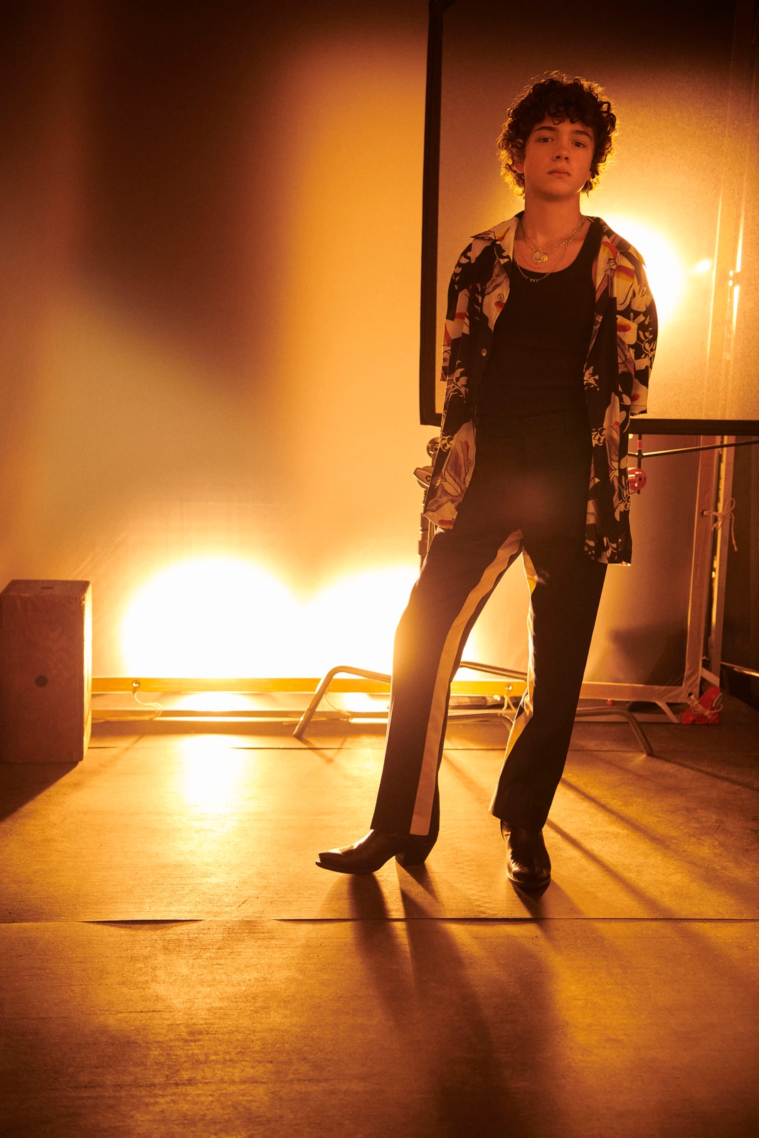 Actor Noah Jupe at TIFF, floral jacket over black tank and stripe trousers, two hot tungsten lights blazing gold behind him.
