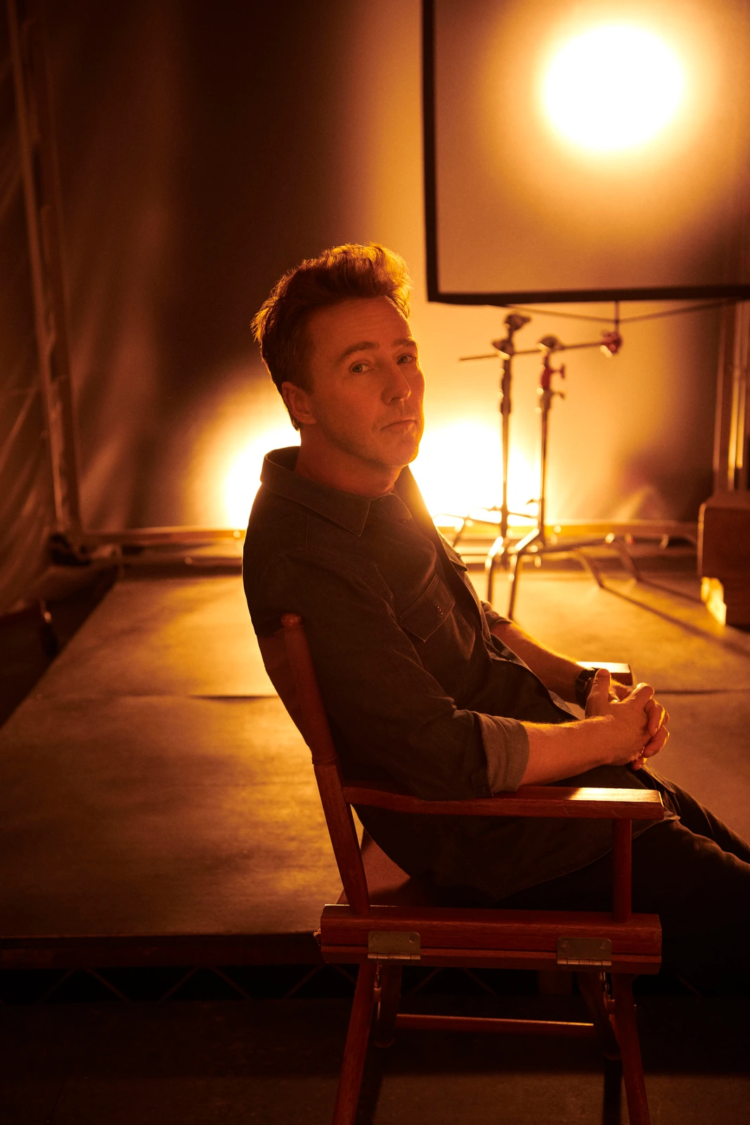 Actor Edward Norton seated in a director's chair, turning over his shoulder in amber studio light, warm glow of a tungsten.