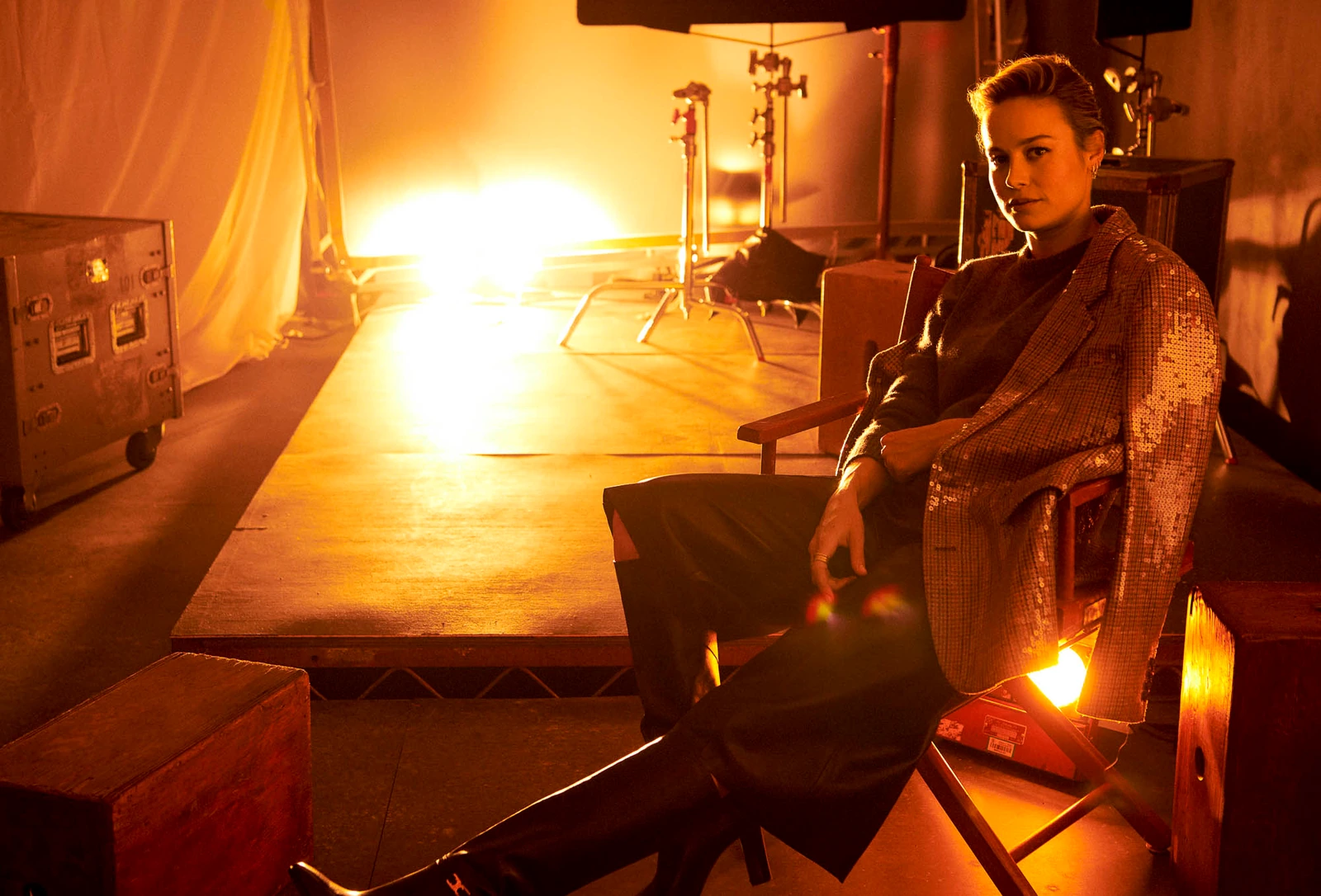 Actor Brie Larson seated in a director's chair on a film stage, sequined blazer catching amber light, gaze cool and steady.