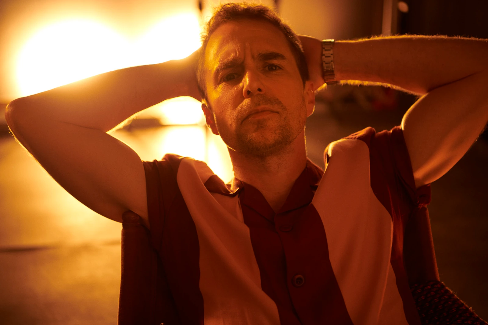 Actor Sam Rockwell, hands behind his head, in a bold burgundy-striped sleeveless shirt, bathed in amber studio light.