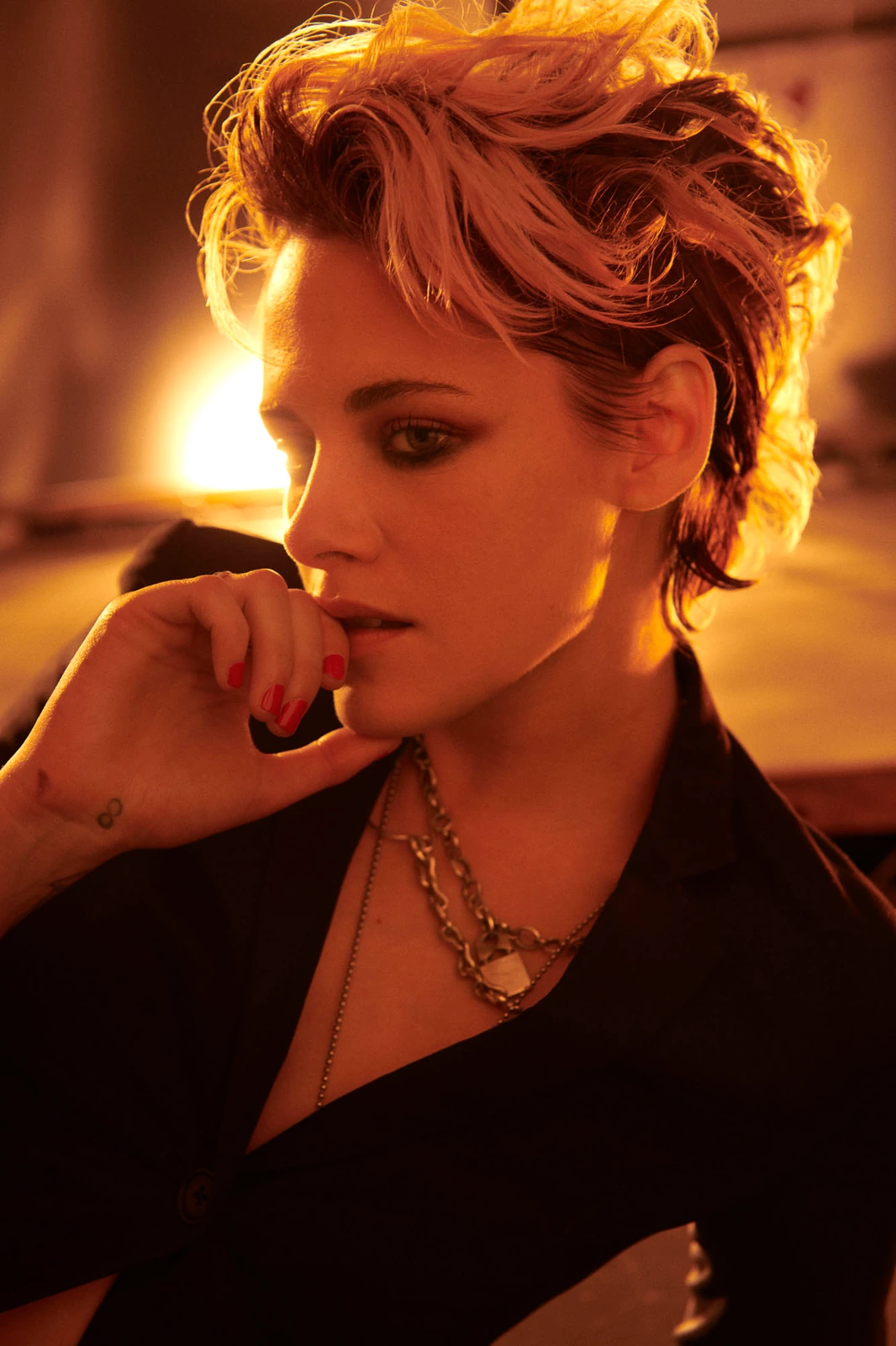 Actor Kristen Stewart, gaze downcast in amber candlelight, tousled short hair, layered chain necklace, red nails at her.
