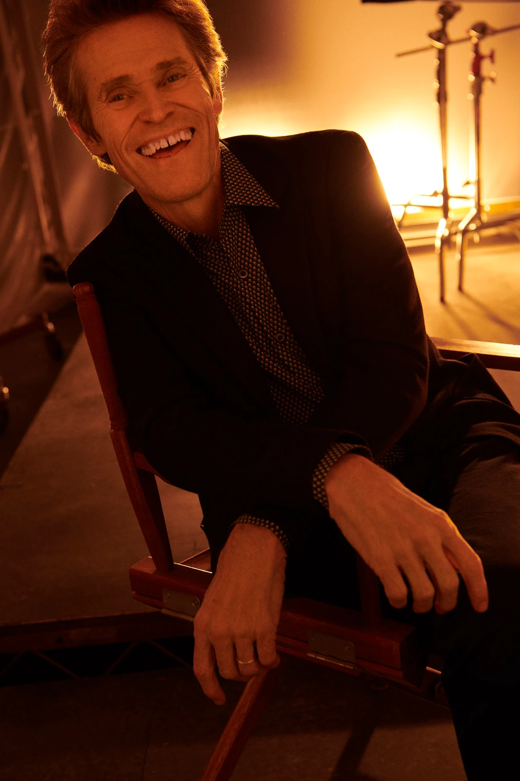 Actor Willem Dafoe laughing openly in a black blazer and dotted shirt, leaning back in a director's chair, warm amber studio.