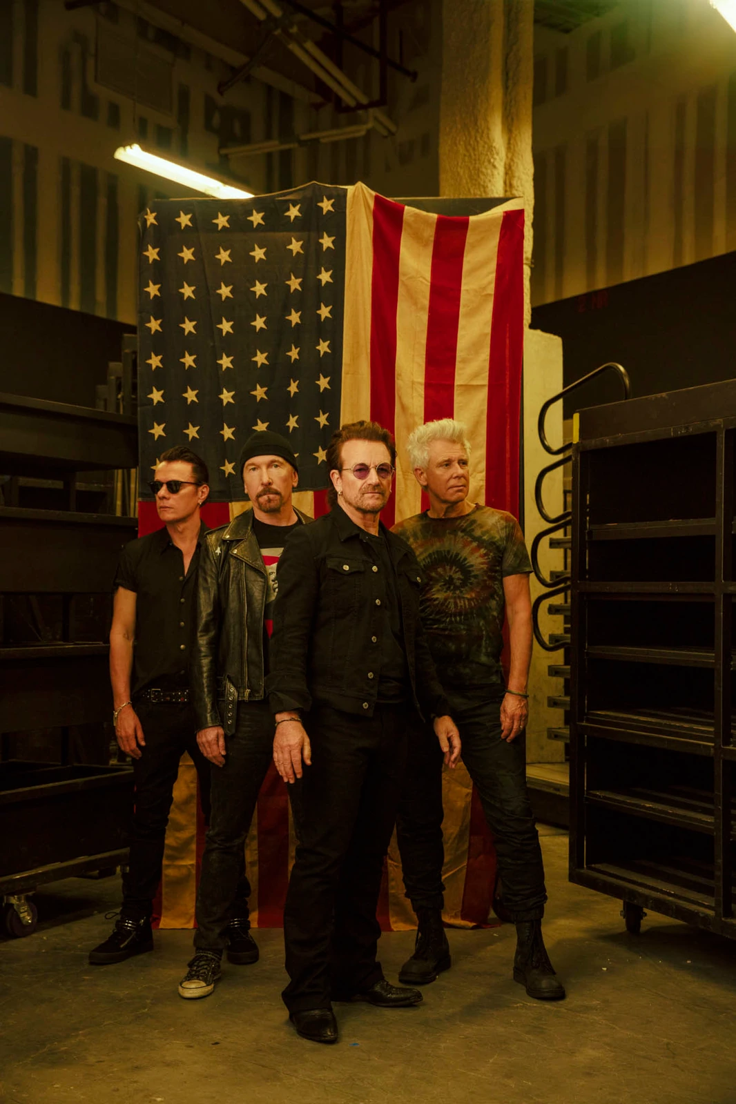 Rock band U2 standing before a worn American flag in a dim backstage space, all black with gritty cool, by Austin Hargrave.