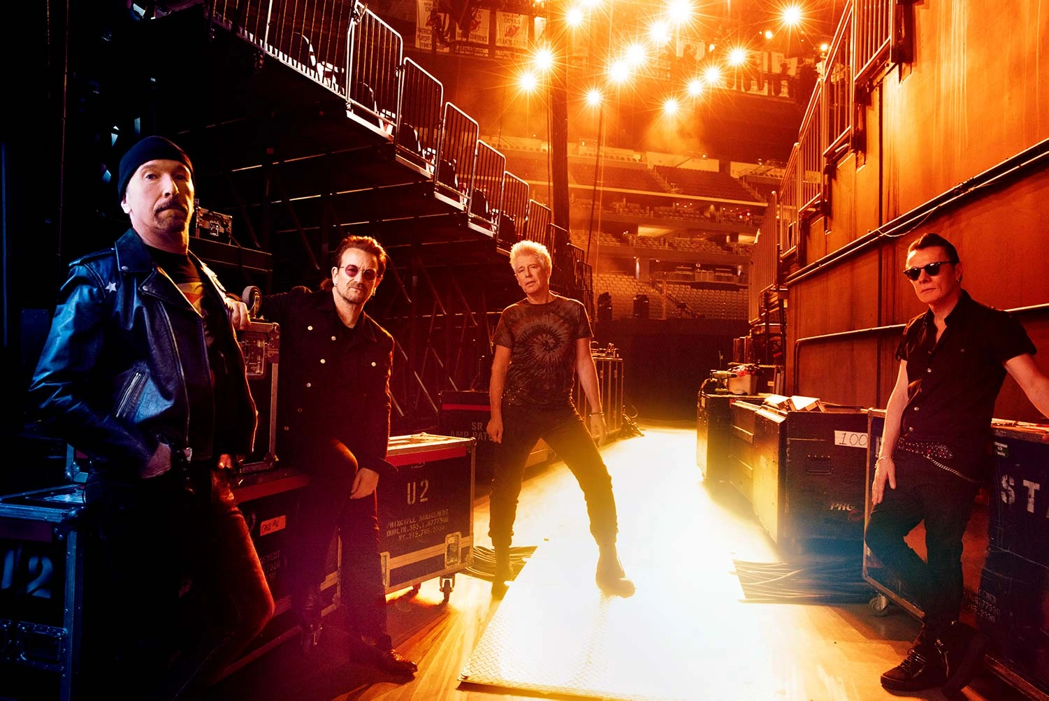 U2 — The Edge, Bono, Larry Mullen Jr. and Adam Clayton — backstage in amber stage light amid road cases, by Austin Hargrave.