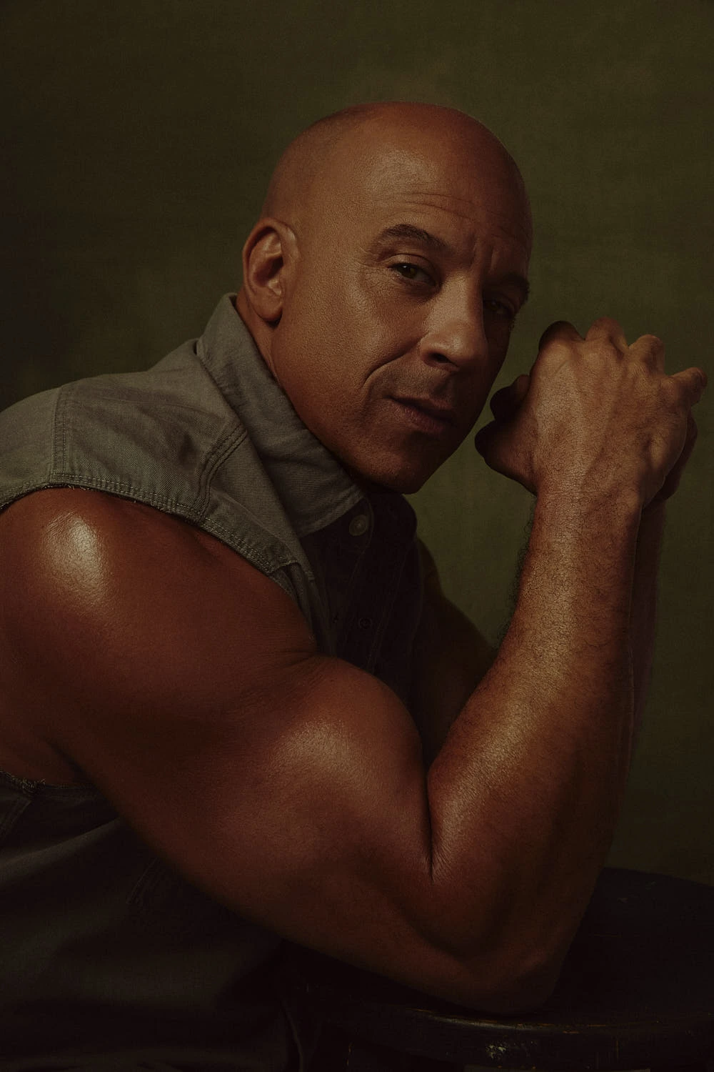 Actor Vin Diesel in a sleeveless denim shirt, arm raised and fist to jaw, warm amber light sculpting muscle against olive.