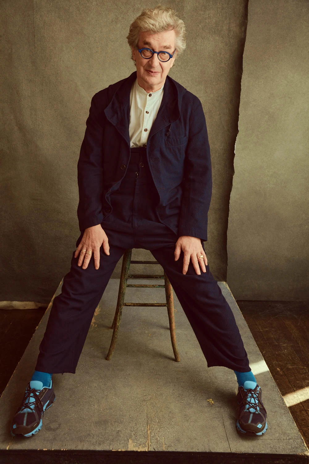 Director Wim Wenders seated wide-legged on a wooden stool, navy jacket and bold blue socks, round glasses, against warm.