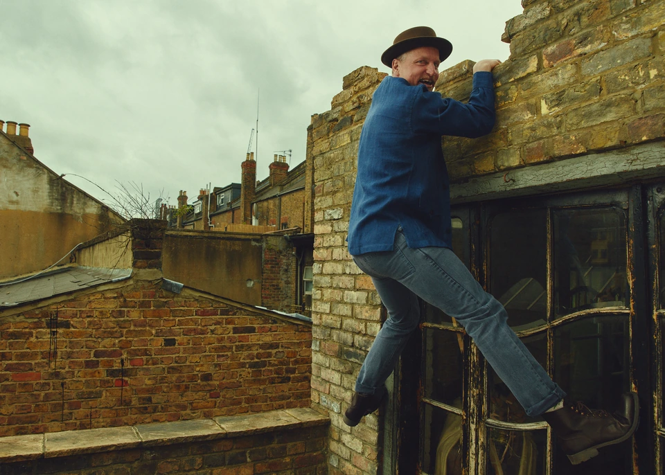 Actor Woody Harrelson scaling a crumbling London rooftop in a blue jacket and brown hat, laughing mid-climb.