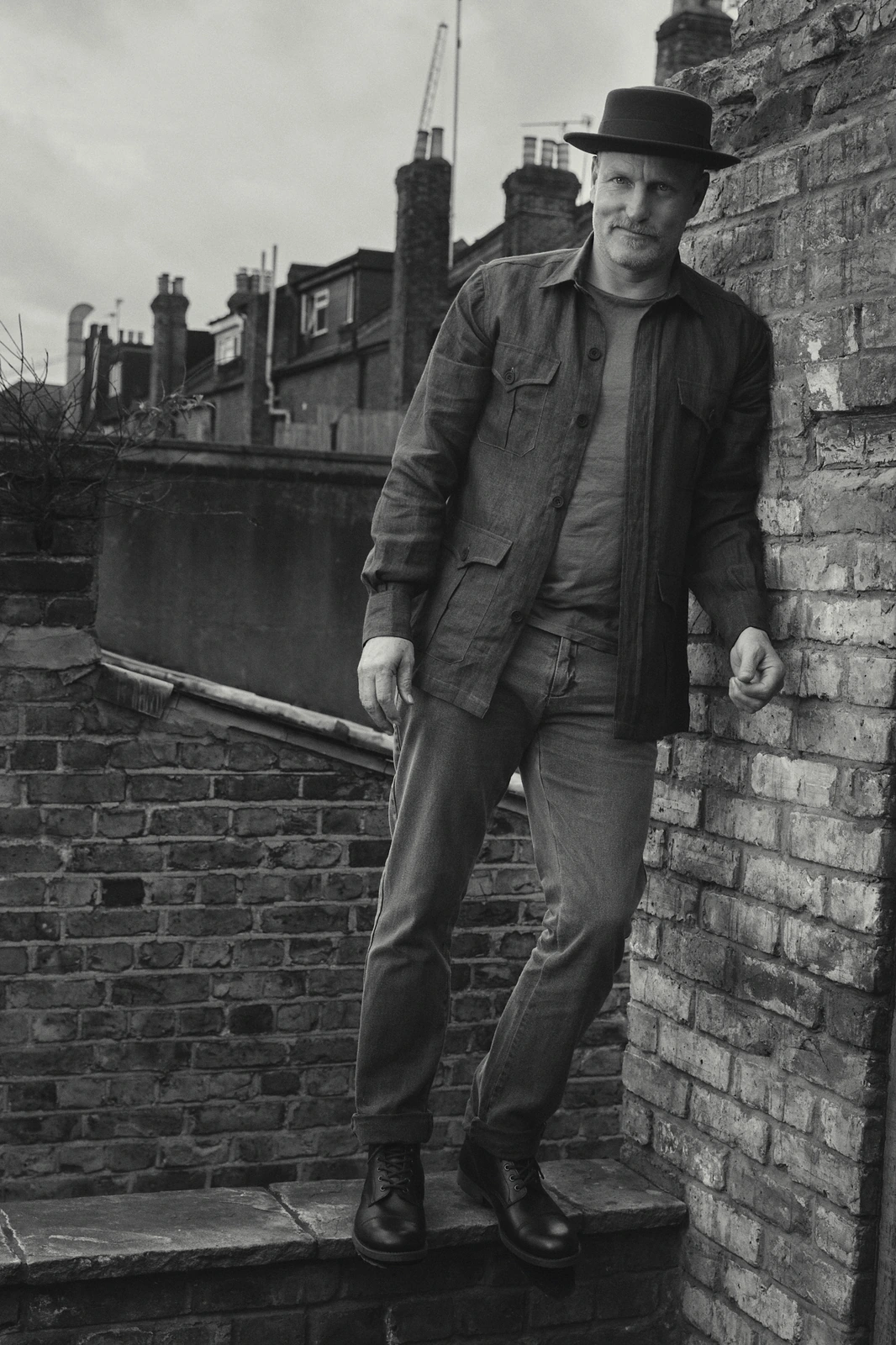 Actor Woody Harrelson leaning on a London brick wall in a flat-brim hat, field jacket, and boots, overcast rooftops behind.