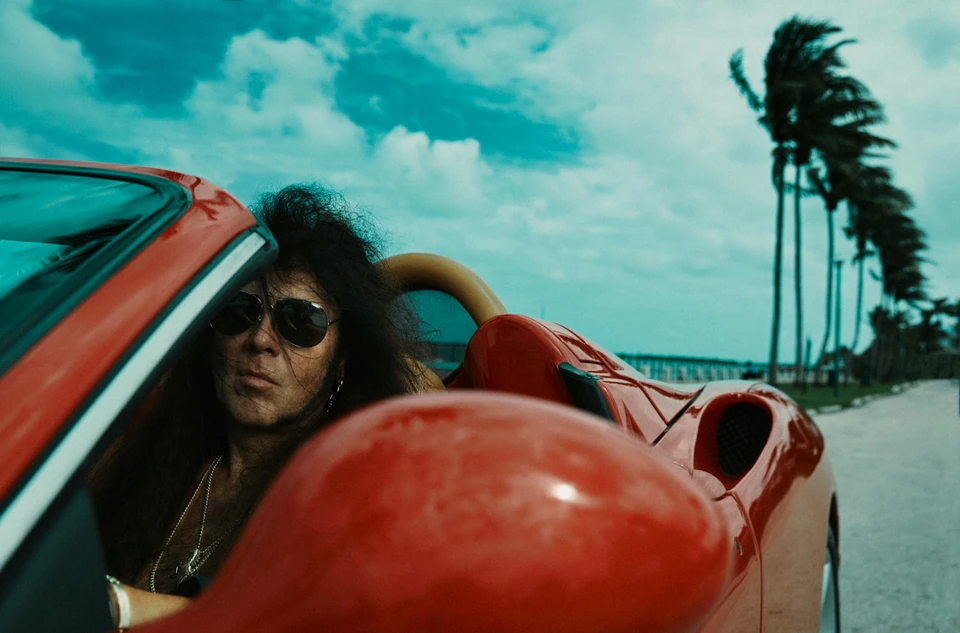 Guitarist Yngwie Malmsteen, wild dark hair and aviators, leaning from a red convertible as palm trees whip in coastal wind.