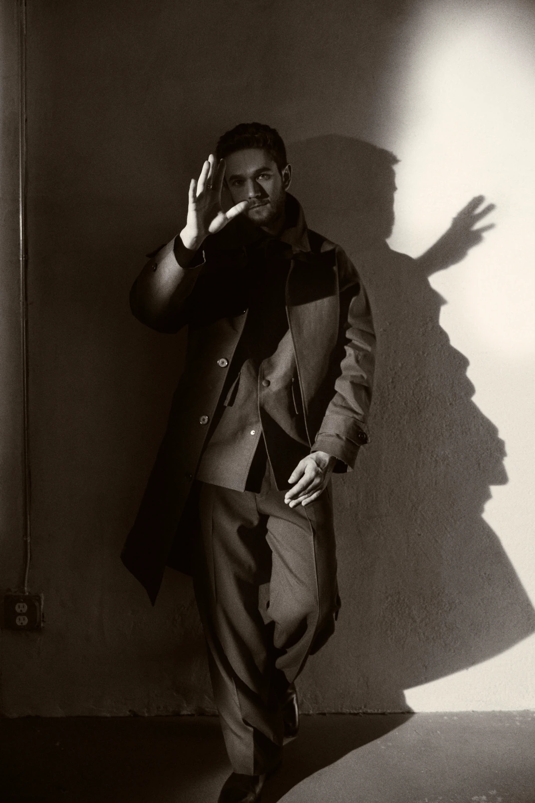 Electronic music producer Zedd leaning against a bare wall, one hand raised, dramatic shadow cast beside him in stark black.