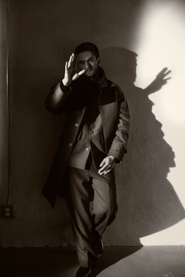 Electronic music producer Zedd leaning against a bare wall, one hand raised, dramatic shadow cast beside him in stark black.